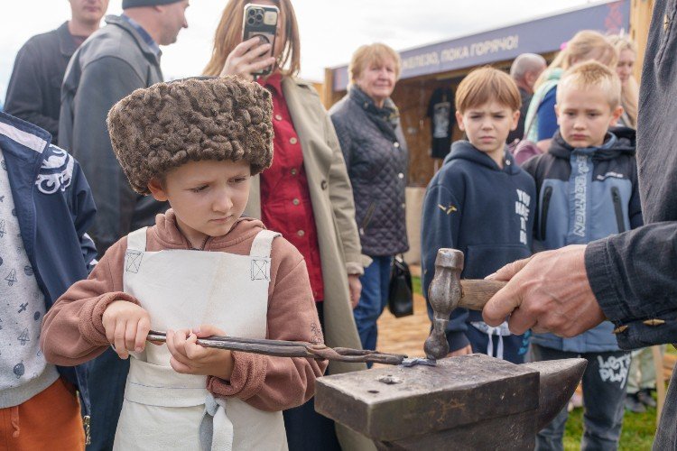 Маленький мальчик в казачьей шапке осваивает азы ковальского искусства