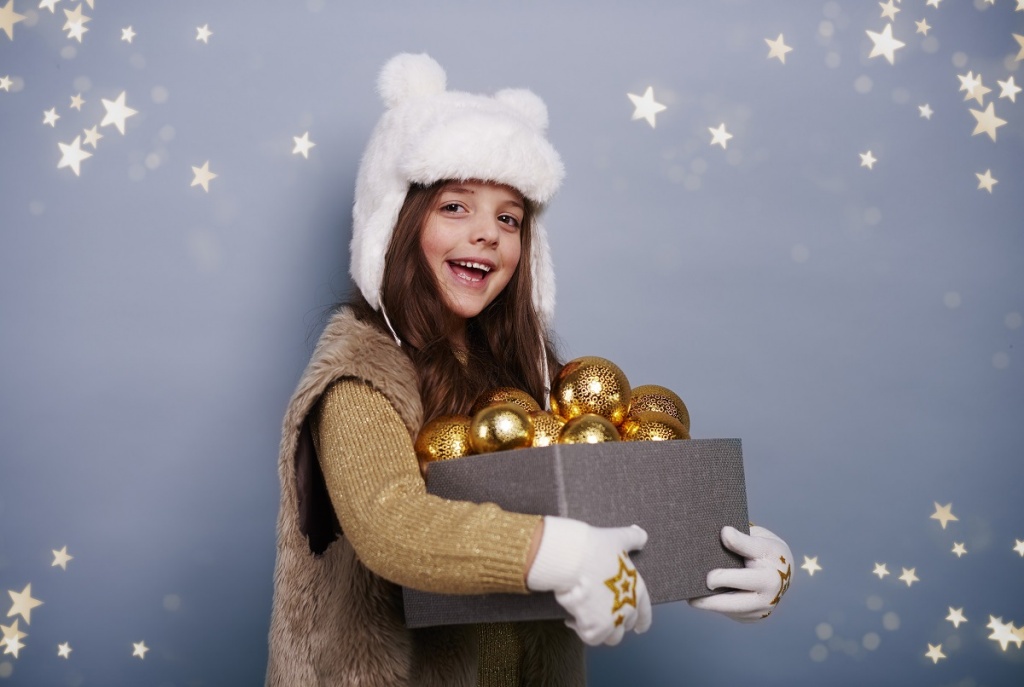 Девочка в белой шапке и варежках держит коробку с новогодними шарами