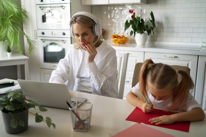Маленькая девочка рисует, сидя за столом рядом с мамой, работающей за компьютером 