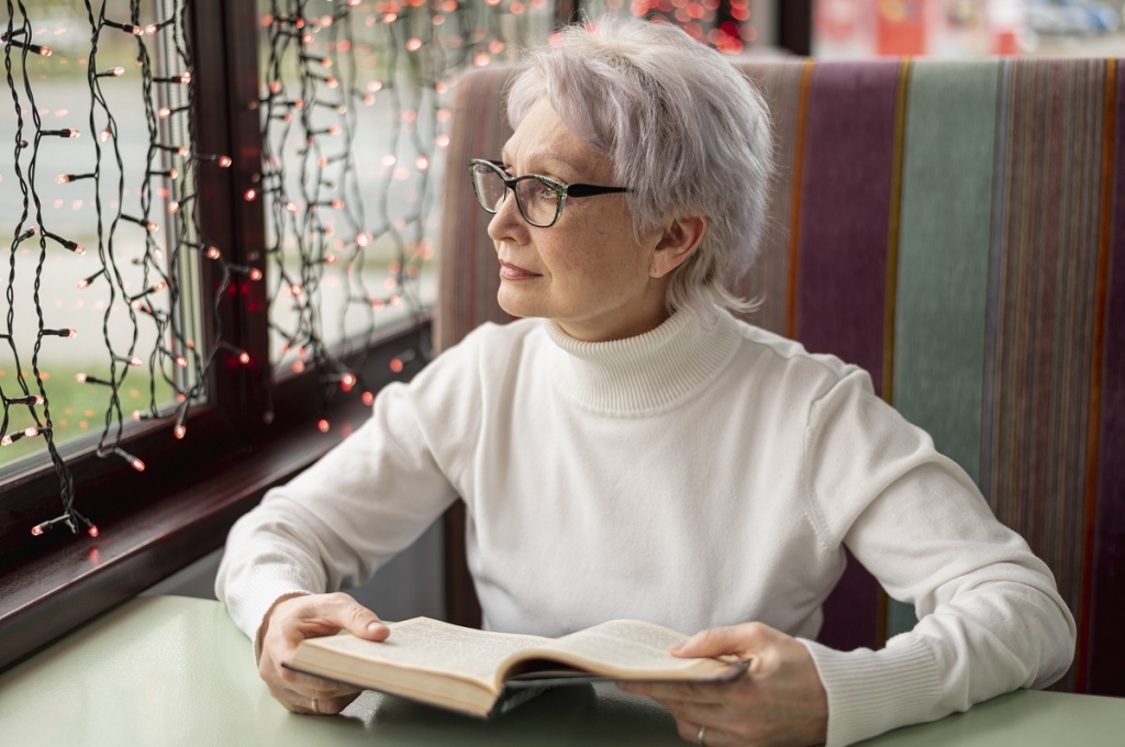 Взрослая женщина сидит с книгой у окна в кафе