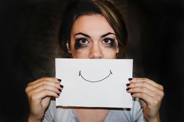 Молодая девушка с размазанной косметикой держит у рта лист бумаги с нарисованной улыбкой