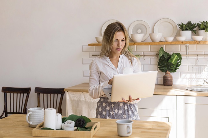 Женщина стоит на кухне в домашней одежде и держит в руках ноутбук