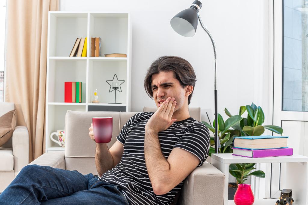 young-man-in-casual-clothes-holding-cup-of-hot-tea-looking-unwell-touching-his-cheek-feeling-toothache-sitting-on-the-chair-in-light-living-room.jpg
