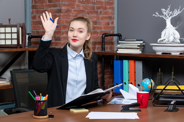 front-view-young-confident-female-assistant-sitting-her-desk-holding-document-saying-hello-someone-office.jpg Девушка здоровается в офисе